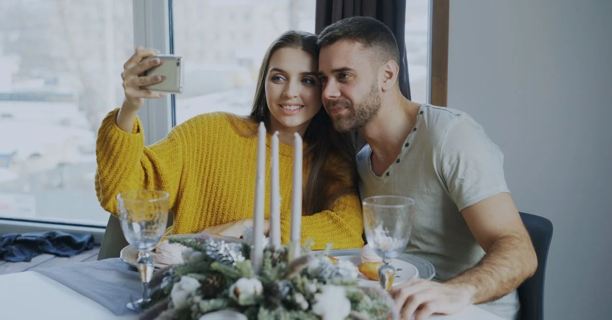 Couple souriant prenant un selfie à une table festive à l'intérieur pendant la saison hivernale.