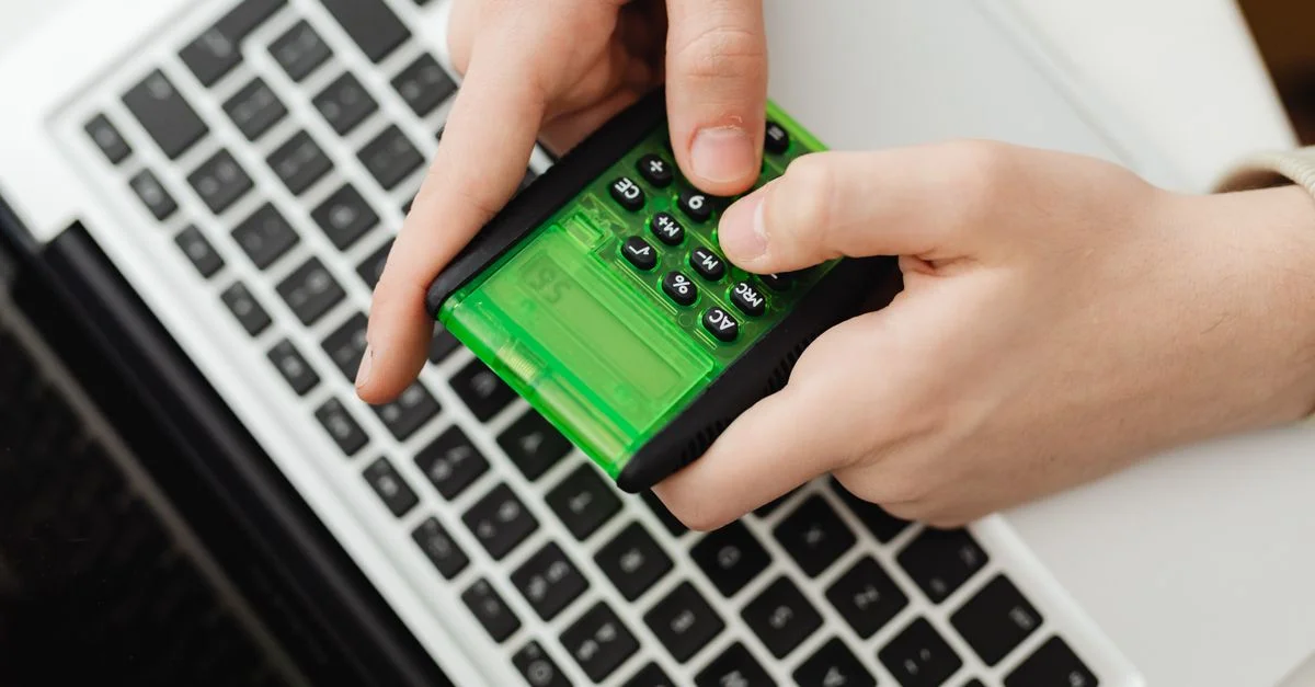 Des mains utilisant une calculatrice verte sur le clavier d'un ordinateur portable, représentant des tâches liées aux affaires et aux finances.