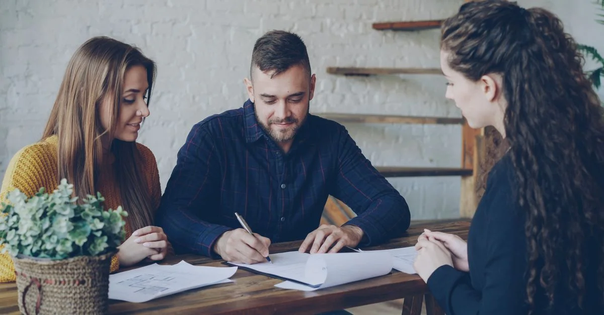 Un jeune couple discutant de documents avec un agent immobilier à l'intérieur.