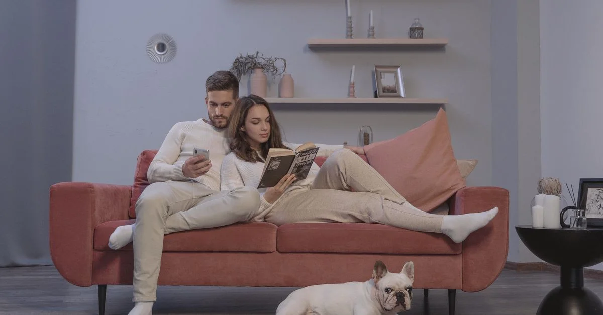 Un couple se détendant sur un canapé rouge, lisant et utilisant un smartphone, avec un bulldog français à proximité.