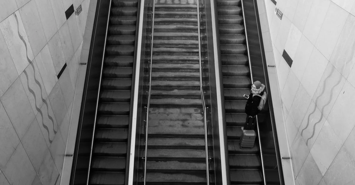 Vue en plongée d'une femme avec une valise sur un escalator dans un cadre urbain.