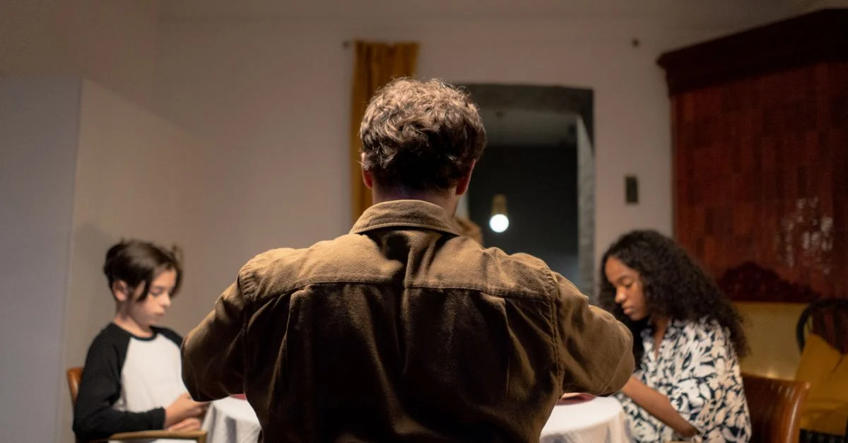 Une famille se regroupe autour d'une table à manger, mettant en scène un père et deux enfants dans un cadre intime.