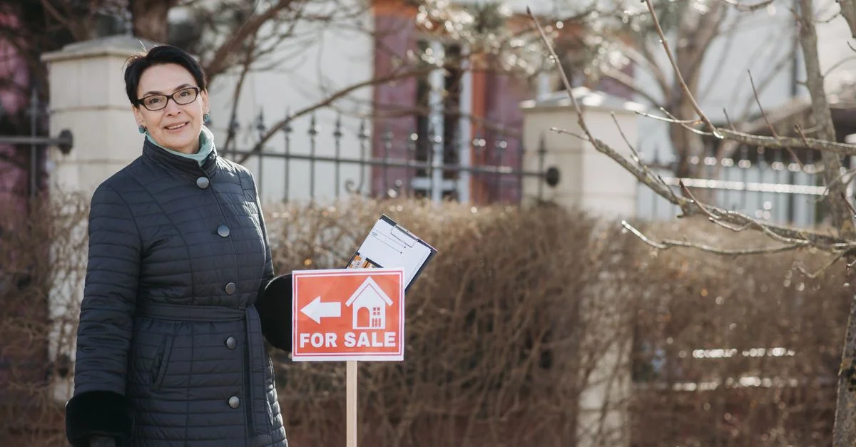 Femme agent immobilier souriante en manteau d'hiver se tenant à côté d'un panneau 'À vendre' avec des documents, à l'extérieur.